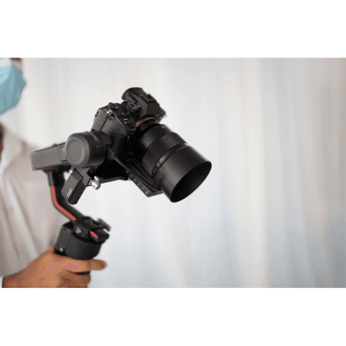 Person holding a camera with a stabilizing gimbal against a blurred white background.